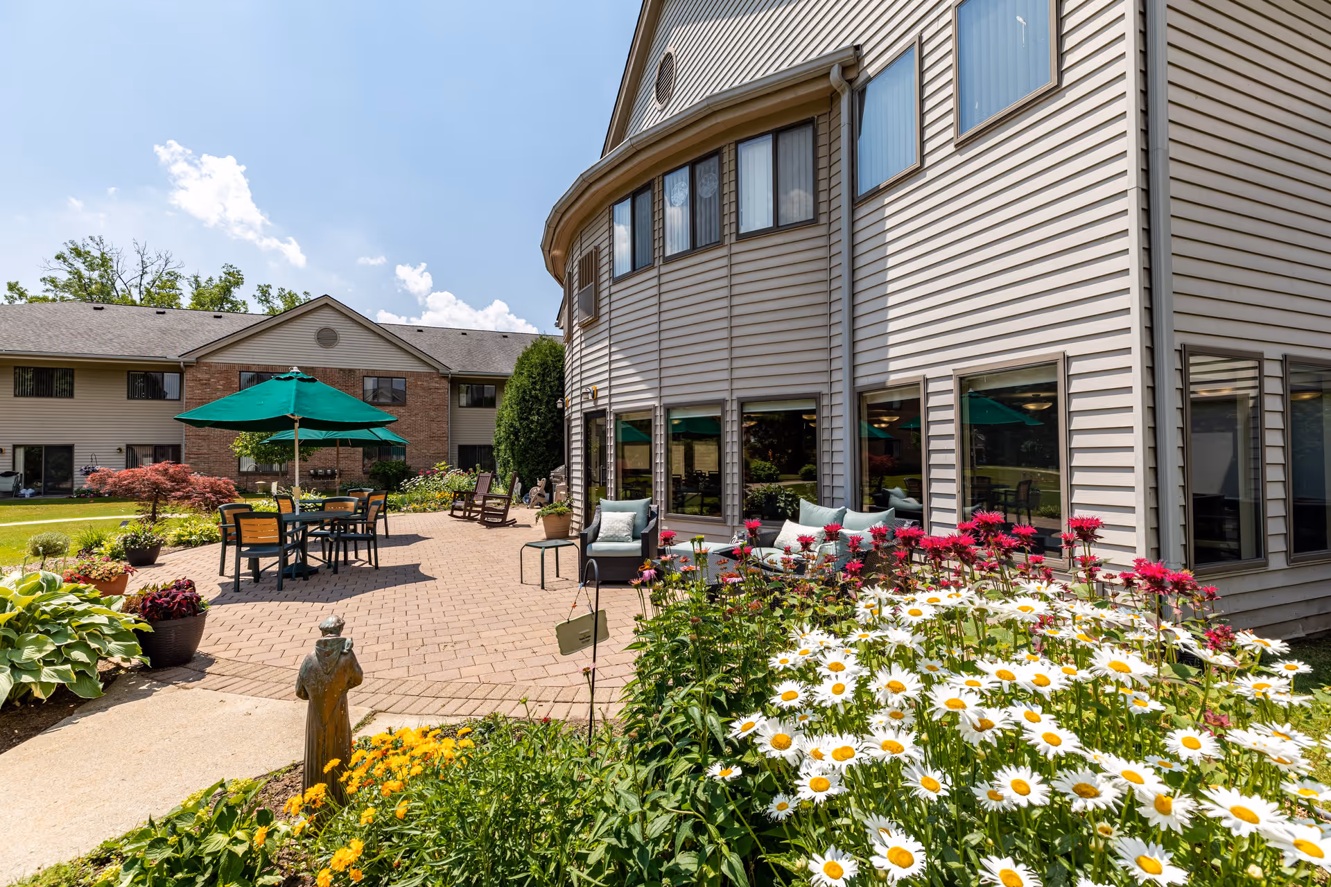 Outdoor patio area at Lourdes Senior Community featuring a paved seating area with tables, chairs, and green umbrellas. The patio is surrounded by colorful flower beds with daisies and other flowers, and the building exterior is visible with large windows. The sky is clear with a few clouds.