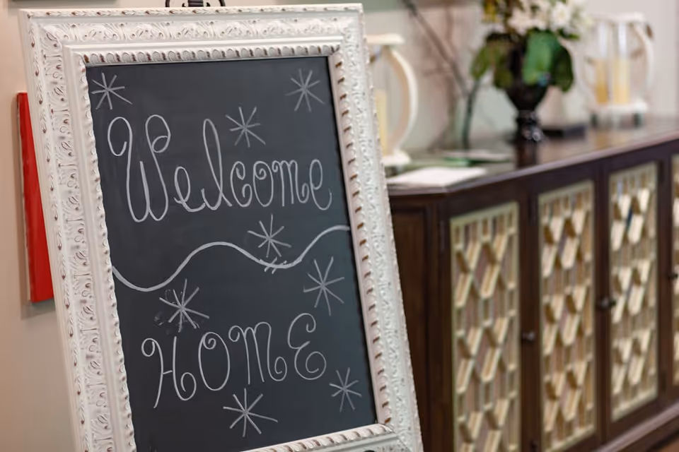 A decorative chalkboard sign with a white ornate frame displaying the handwritten message 'Welcome HOME' surrounded by star-like drawings, placed indoors next to a wooden cabinet with patterned doors and some decorative items on top.