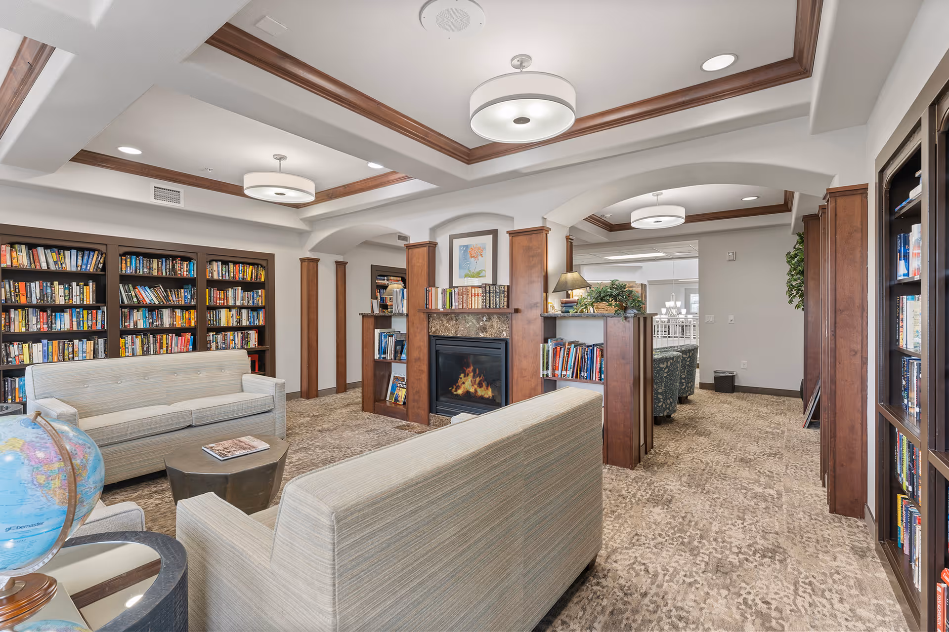 Communal library lounge with bookshelves, upholstered sofas, a fireplace, and overhead lighting.
