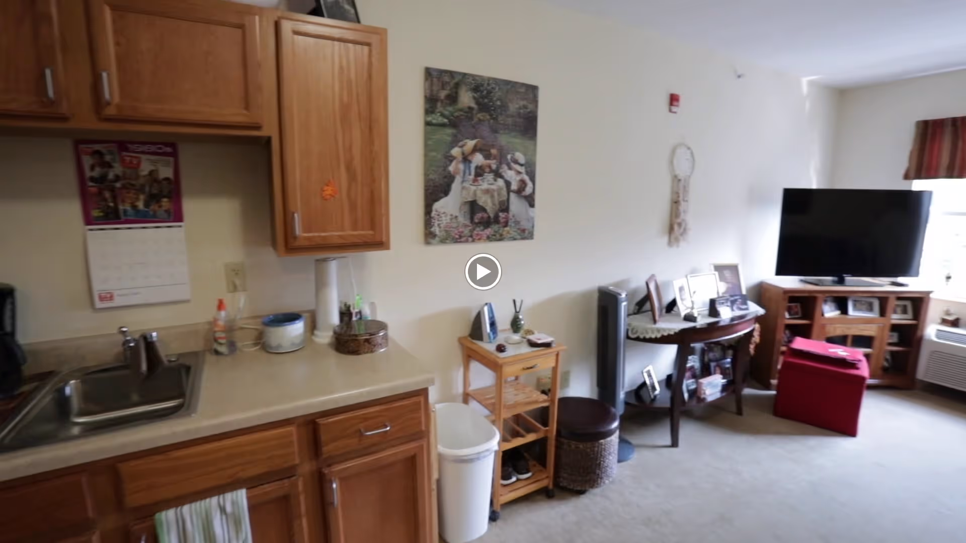 Interior view of a senior living facility room showing a small kitchenette area with a sink, countertop, and wooden cabinets on the left. Adjacent to the kitchenette is a small wooden shelving unit with various items on top and a trash can below. The room extends into a living area with a TV on a wooden stand, a red ottoman, a small table with framed photos, and a window with a valance. The walls are decorated with a painting and a hanging ornament.