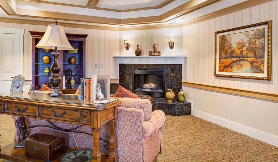 A cozy living room area with a wooden desk holding a lamp, books, and decorative items in the foreground. Behind the desk is a comfortable armchair facing a black marble fireplace with a fire burning. On the wall above the fireplace are decorative vases and two wall sconces. To the right, a framed painting of a serene autumn landscape hangs on the wall. The room has beige wallpaper with a subtle pattern and a carpeted floor.