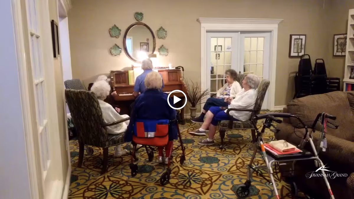 Several elderly residents seated in a common room around a piano while one person plays, with chairs and walkers visible.