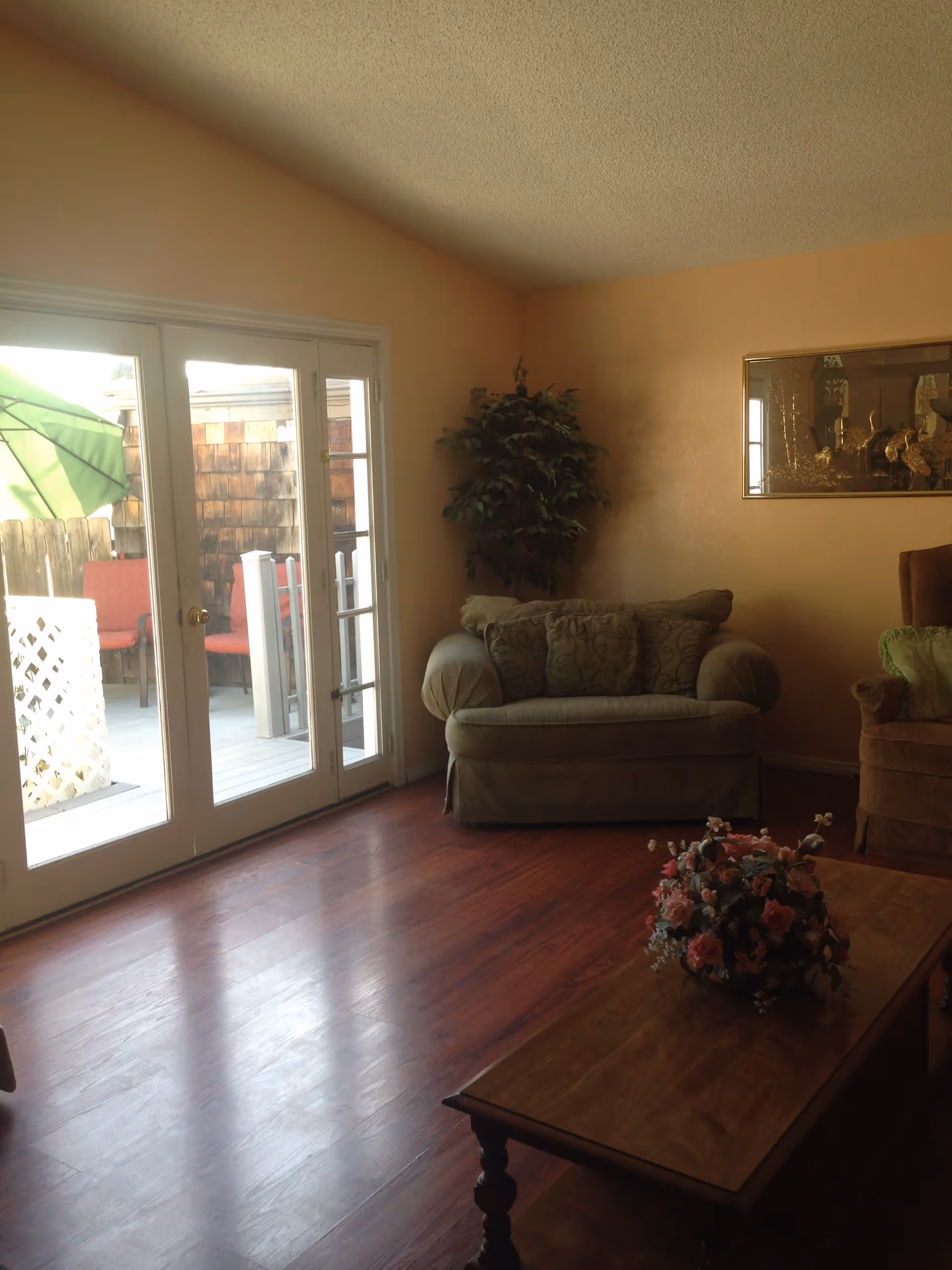 A cozy living room with wooden flooring, a cushioned armchair, a loveseat with patterned pillows, a wooden coffee table with a floral arrangement, a potted plant in the corner, and glass doors leading to an outdoor patio with red chairs and a green umbrella.