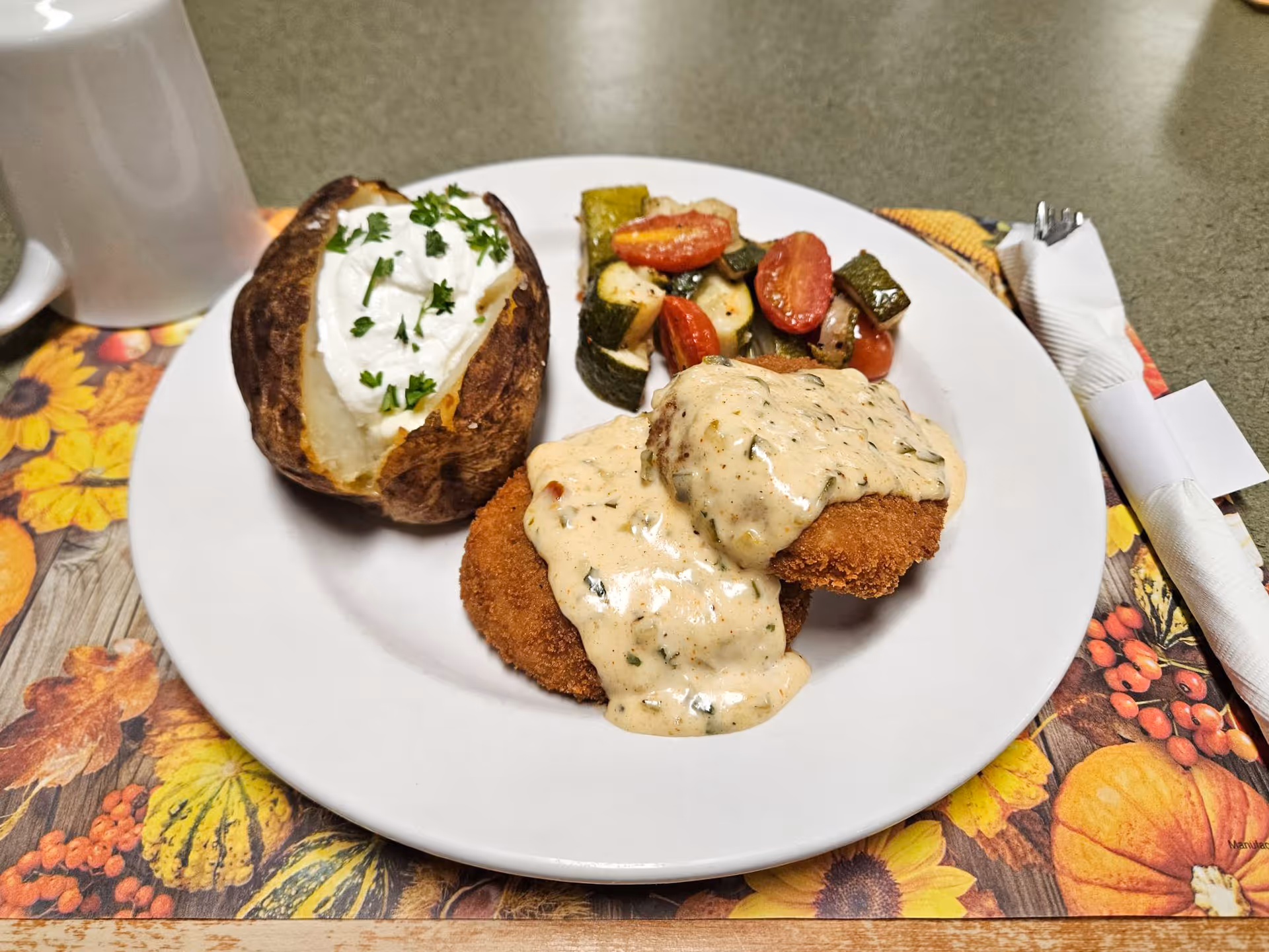 A plate with a baked potato topped with sour cream and parsley, two breaded patties covered in creamy sauce, roasted zucchini and cherry tomatoes, plus a coffee mug and wrapped cutlery on a decorative placemat.