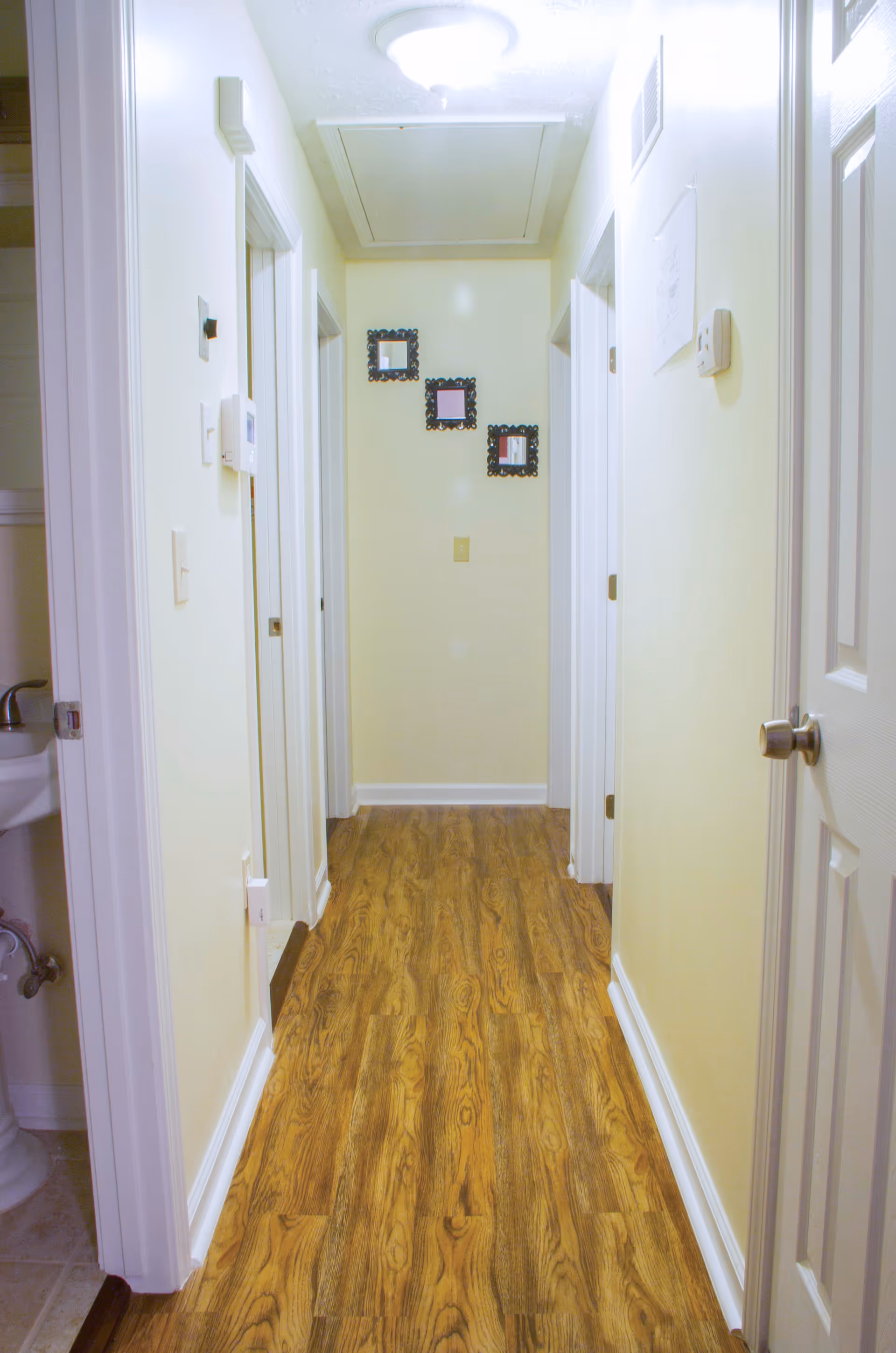 A narrow hallway with light yellow walls and wood-patterned flooring. There are three small decorative framed mirrors on the far wall. Several white doors line the hallway on both sides, and a ceiling light fixture is visible. A small sink is partially visible through an open door on the left.