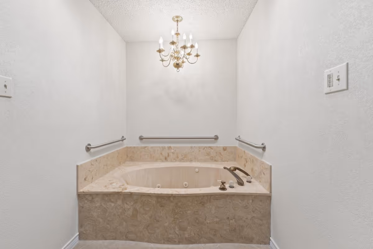 A beige marble bathtub with built-in jets is set against a white wall with three metal grab bars. Above the tub hangs a gold chandelier with multiple candle-style lights. The walls and ceiling are white, and there are two light switches on the walls near the tub.