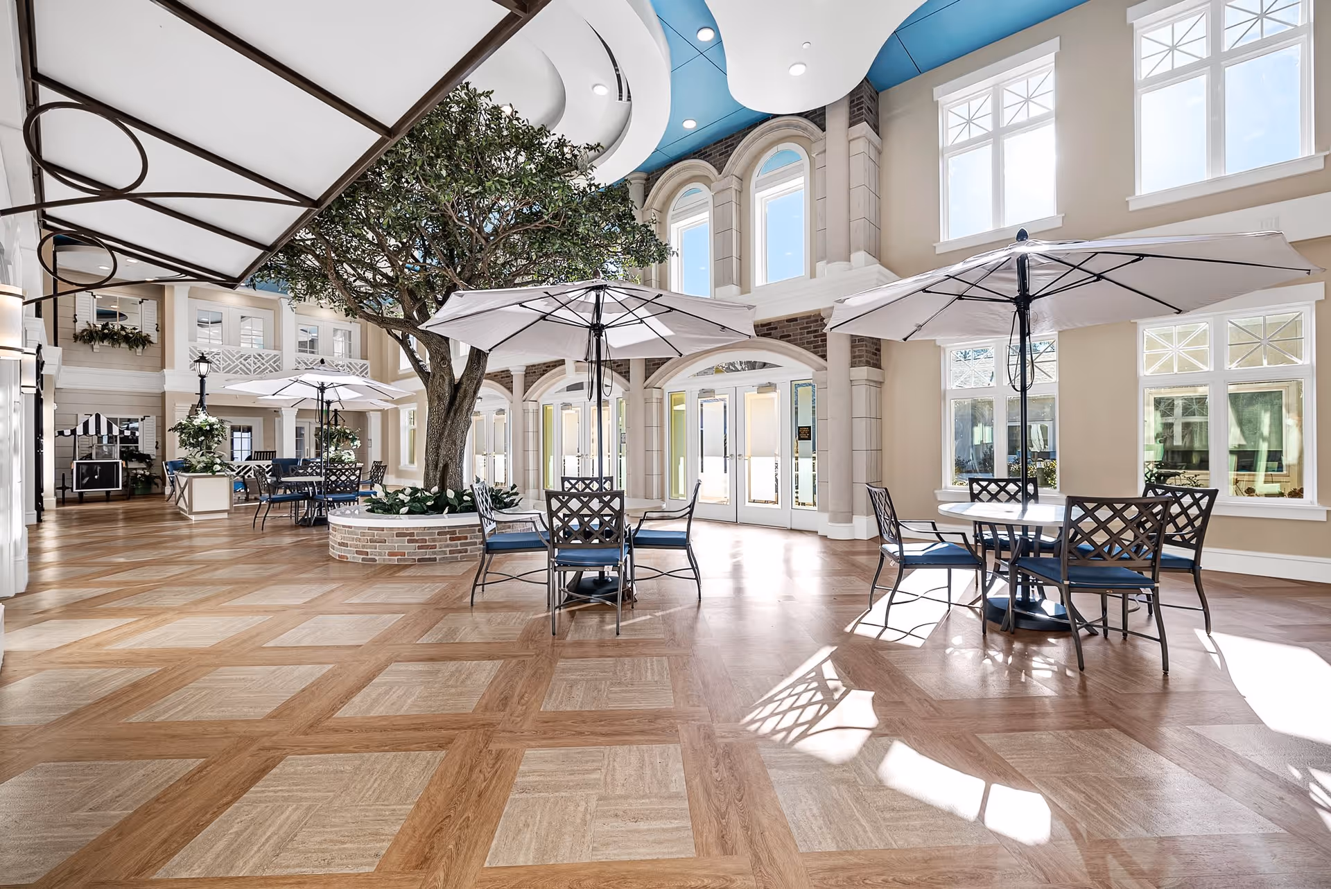Bright indoor common area with large windows letting in natural light, featuring several round tables with chairs and large white umbrellas. A large tree is planted in a circular brick planter in the center of the space. The floor has a patterned wood design, and the ceiling has a blue and white decorative element.