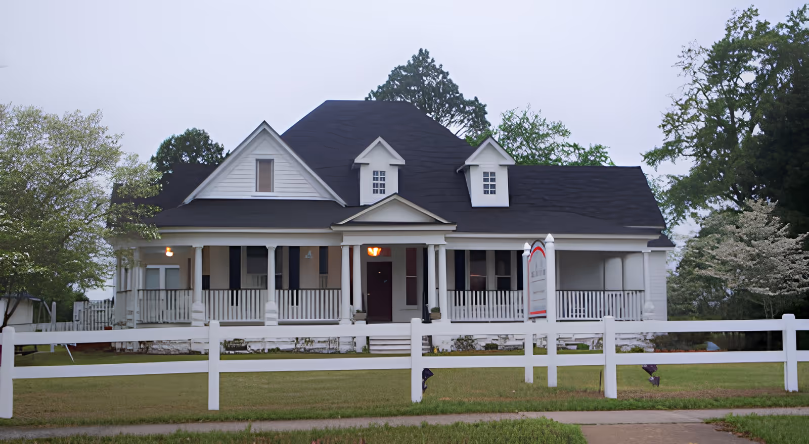 Front exterior view of a large white house with a black roof, multiple windows, a porch with white railings, and a white fence in front. There are trees on both sides and a signpost near the sidewalk.