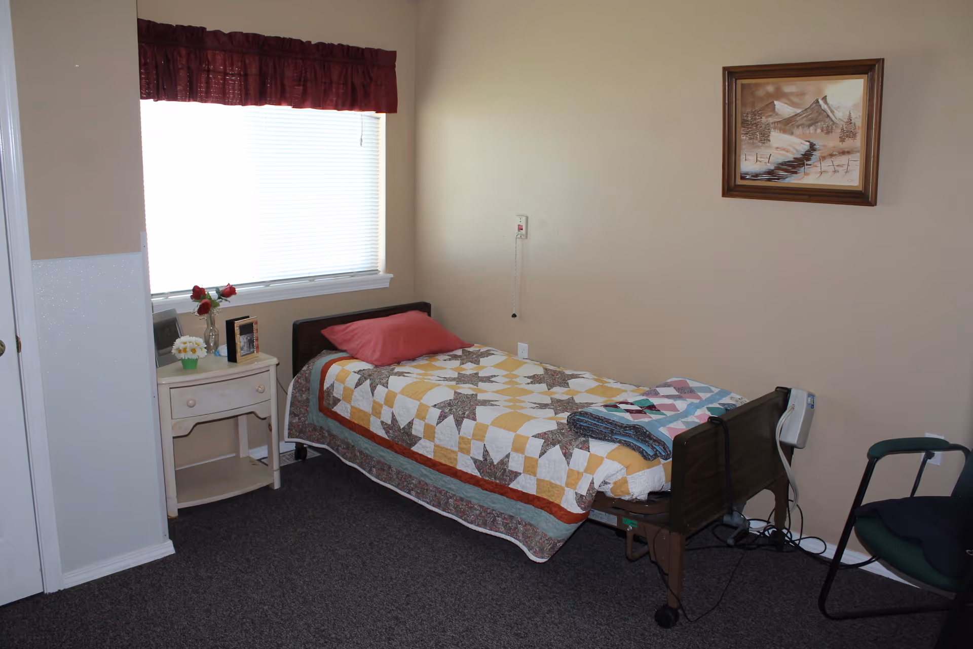 A small bedroom with a single bed covered by a colorful quilt and a red pillow. Next to the bed is a white nightstand with a vase of red roses, a small potted plant, and a framed photo. A window with closed blinds and a red valance is above the nightstand. On the wall above the bed is a framed painting of a mountain landscape. There is a green chair with black armrests near the foot of the bed. The room has beige walls and dark carpeted flooring.