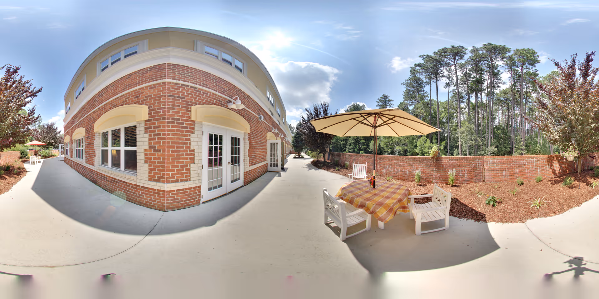 Outdoor patio area at Fox Hollow Senior Living Community featuring a brick building with multiple windows and doors. The patio has a table covered with a checkered tablecloth and an umbrella, surrounded by white chairs. There are trees and landscaped plants along a brick wall under a partly cloudy sky.