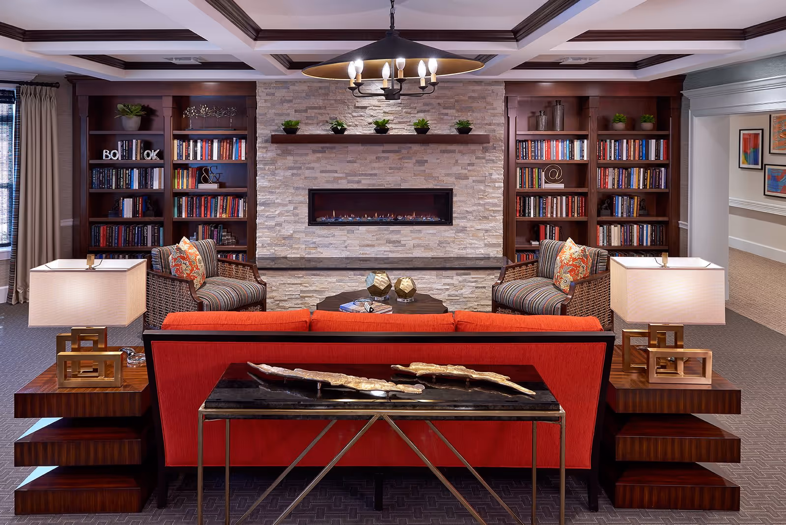 A cozy living room area with a red sofa facing a modern stone fireplace. On either side of the fireplace are built-in wooden bookshelves filled with books and decorative items. Two striped armchairs with patterned cushions are positioned near the fireplace. Two matching table lamps with square shades sit on wooden side tables flanking the sofa. A black console table with decorative pieces is placed behind the sofa. The room has a warm and inviting atmosphere with neutral walls and carpeted flooring.
