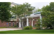 Brick two-story nursing facility entrance with a covered portico supported by columns, surrounded by trees and lawn.
