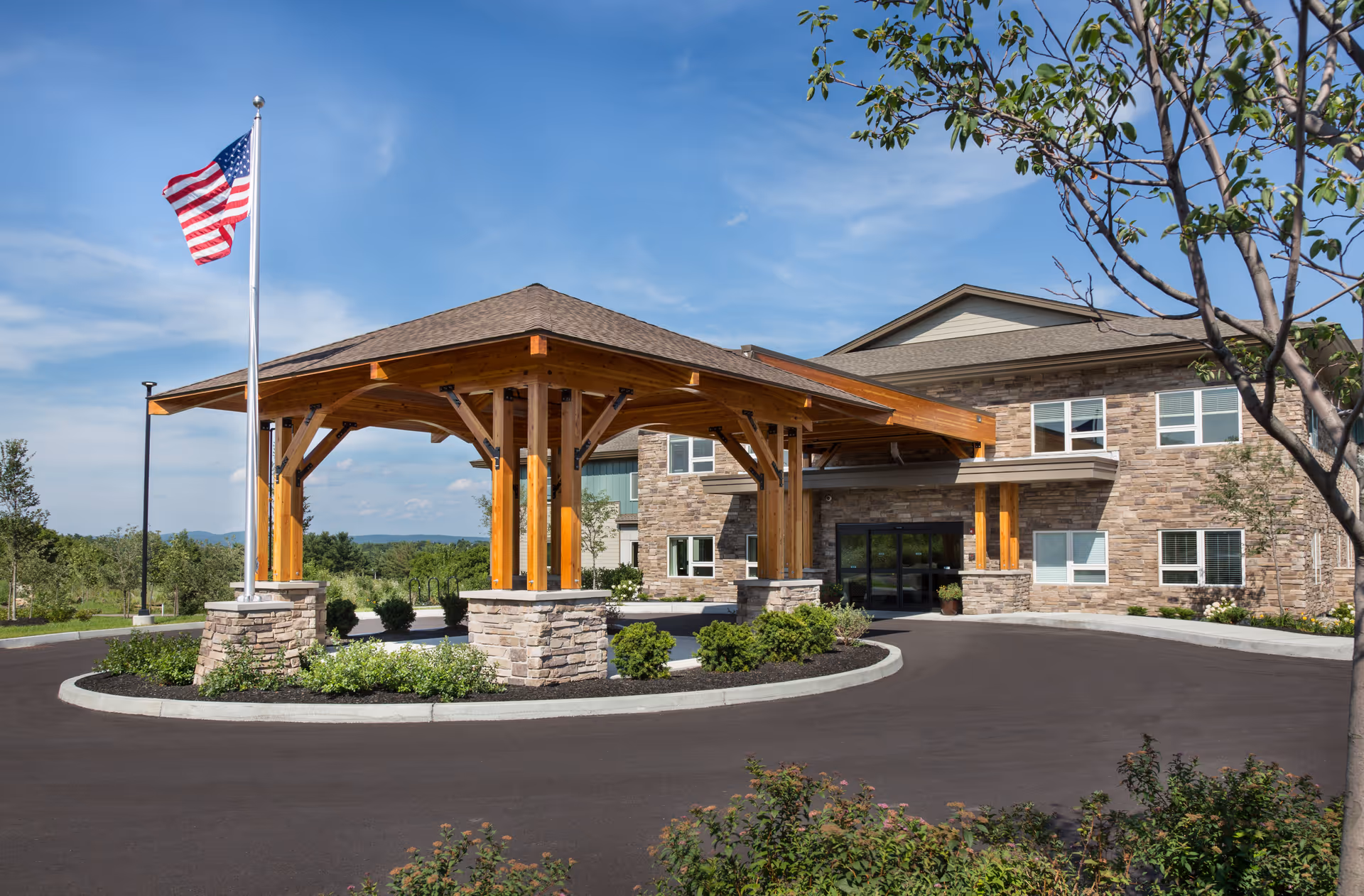 Exterior view of The Residence at Quarry Hill showing a covered entrance with wooden beams and stone pillars, an American flag on a flagpole, and a two-story building with stone and siding under a blue sky.