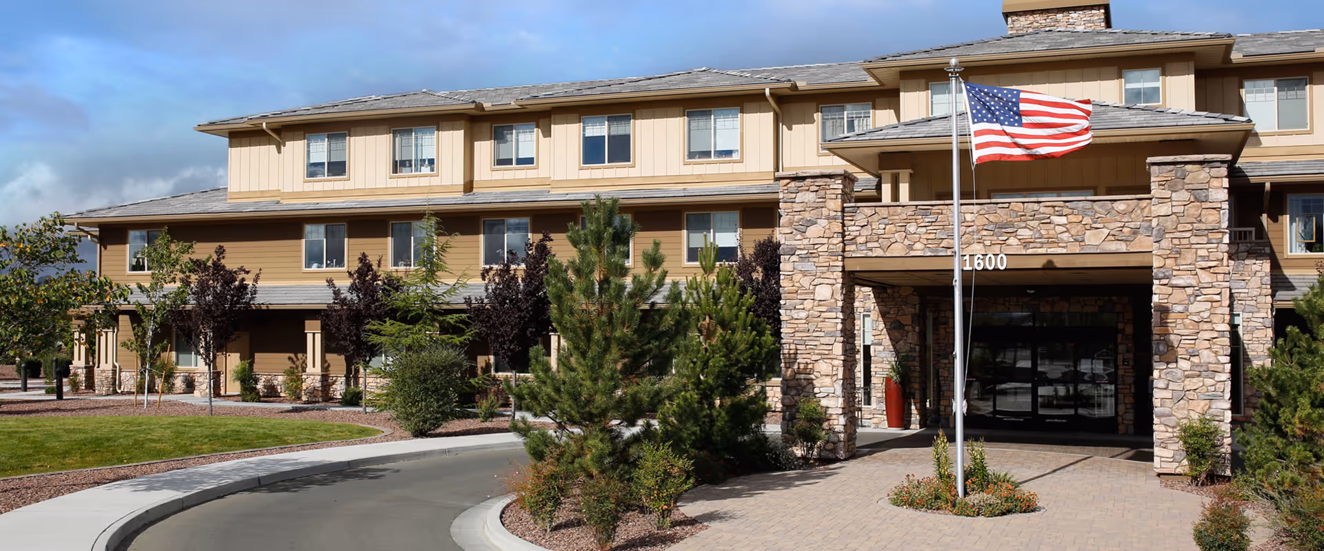 Front exterior of a multi-story senior living building with a stone entrance, American flag, landscaping, and a curved driveway.