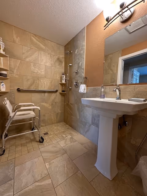 A bathroom with beige tiled walls and floor, featuring a walk-in shower area with grab bars and a shower chair. There is a white pedestal sink with a mirror above it and a soap dispenser on the sink. The lighting fixture above the mirror has two lights.