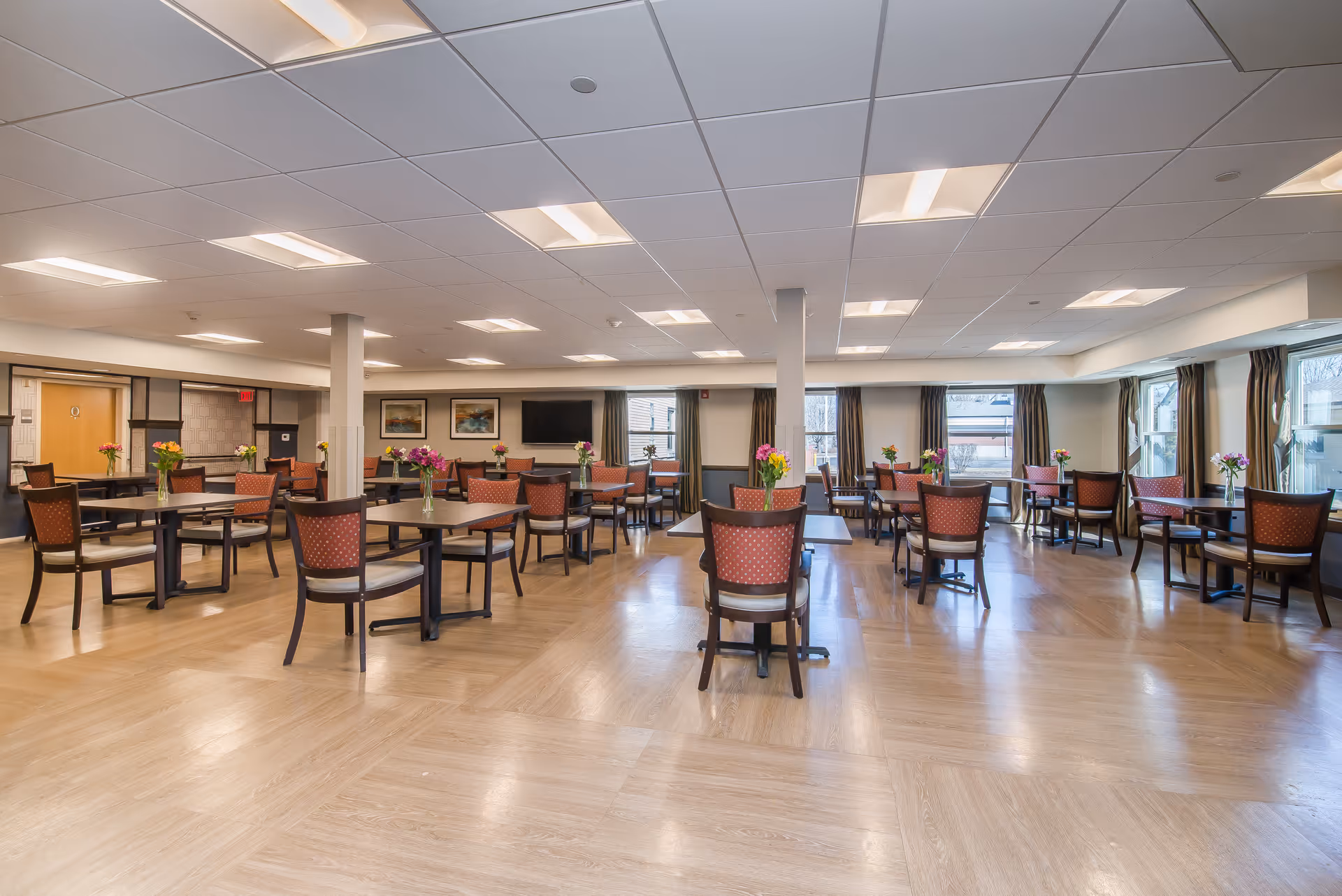 A spacious dining room with multiple round tables and chairs arranged neatly. Each table has a small vase with colorful flowers. The room has large windows with curtains allowing natural light to enter, and the floor is covered with light wood-patterned flooring. The ceiling has recessed lighting panels, and there are framed pictures and a flat-screen TV mounted on the far wall.