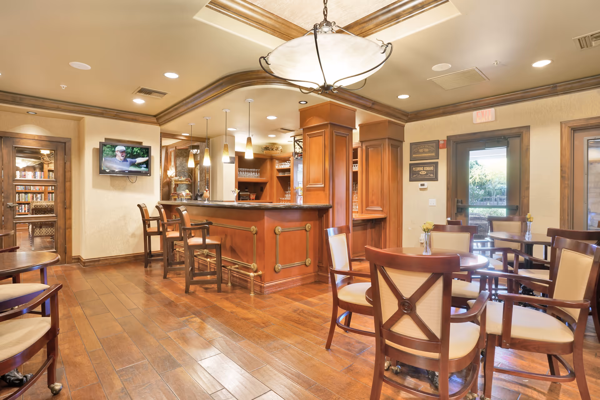 A warm, inviting interior space featuring a wooden bar with high chairs, round tables with cushioned chairs, a ceiling light fixture, and a wall-mounted TV. The room has wooden flooring, beige walls, and large windows and doors letting in natural light. There is a glimpse of a cozy library area through a doorway on the left.