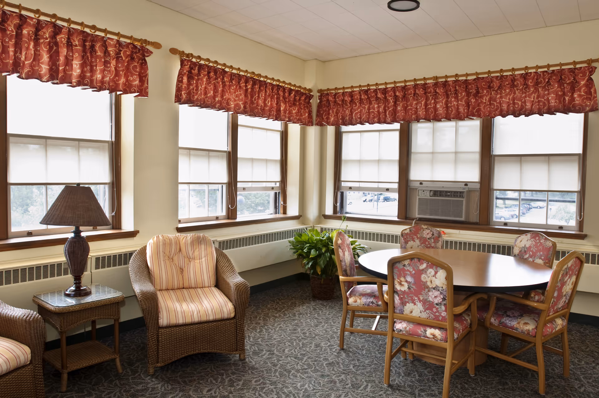 Communal sitting and dining room with wicker armchairs, a round table surrounded by floral-upholstered chairs, and large windows with red valances.