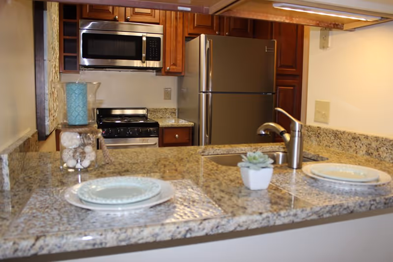 A modern kitchen with granite countertops, a stainless steel refrigerator, microwave, and stove. The countertop has two place settings with plates and a small potted succulent plant near the sink.