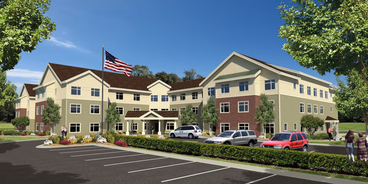 Three-story senior living facility building with a landscaped parking lot, American flag, and a few people near the entrance under a clear blue sky.