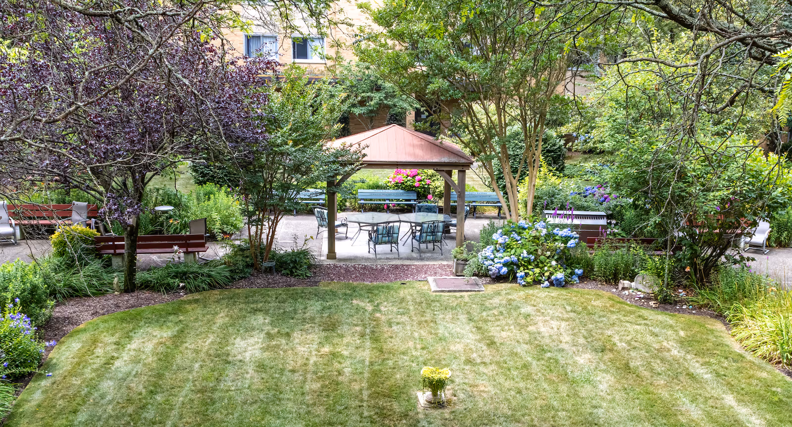 A peaceful outdoor garden area at Maria Regina Residence featuring a well-maintained lawn, various trees and flowering plants, benches along the pathways, and a covered seating area with chairs and a table.