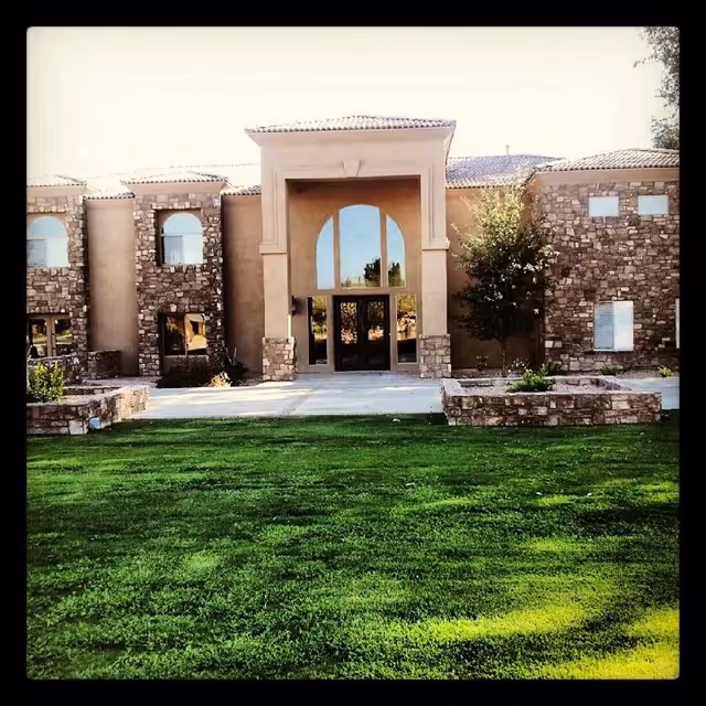 Front exterior view of a senior living facility building with stone and stucco facade, large arched entrance with glass doors, and a well-maintained green lawn in the foreground.
