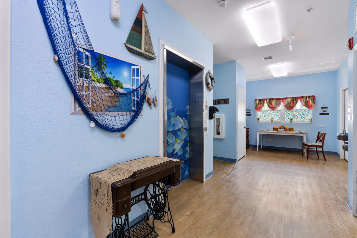 Interior hallway of Marion Oaks Assisted Living with light blue walls, nautical decorations, an elevator and a small table and chair by a window.