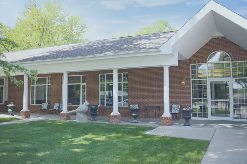 Exterior view of a brick building with white columns supporting a covered porch. There are several chairs and planters on the porch, a large bird sculpture, and a glass door entrance. Green grass and trees surround the building under a partly cloudy sky.