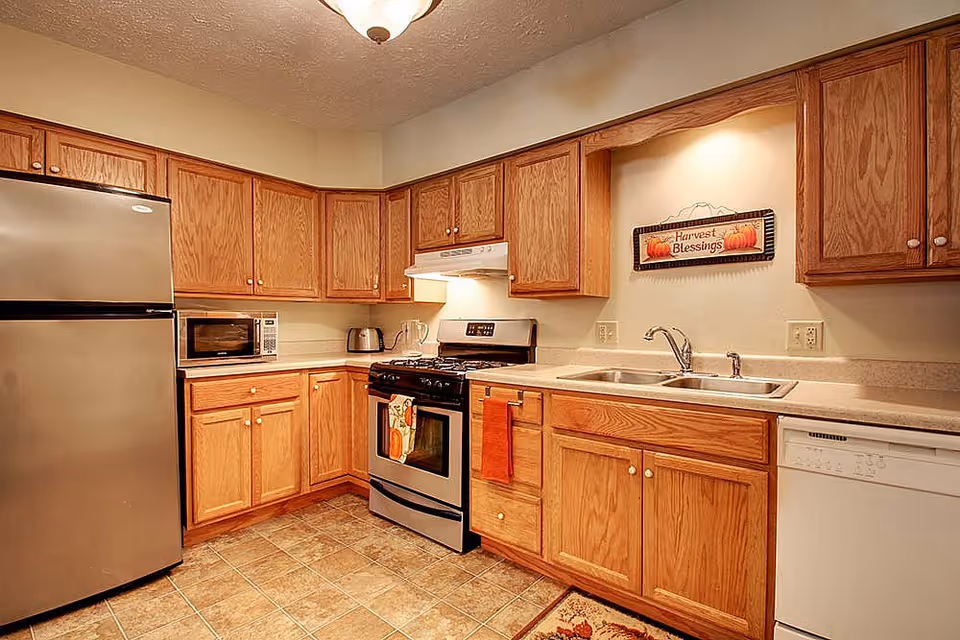 A kitchen with wooden cabinets, a stainless steel refrigerator, a microwave, a toaster, a gas stove with an oven, a double sink, and a dishwasher. There is a decorative sign on the wall above the sink that reads 'Harvest Blessings'. The floor is tiled and there is a ceiling light fixture.