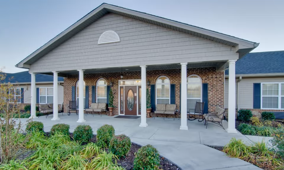 Front exterior view of Willow Springs Senior Living building with a covered porch supported by white columns, outdoor seating, brick and siding walls, and landscaped greenery in the foreground.
