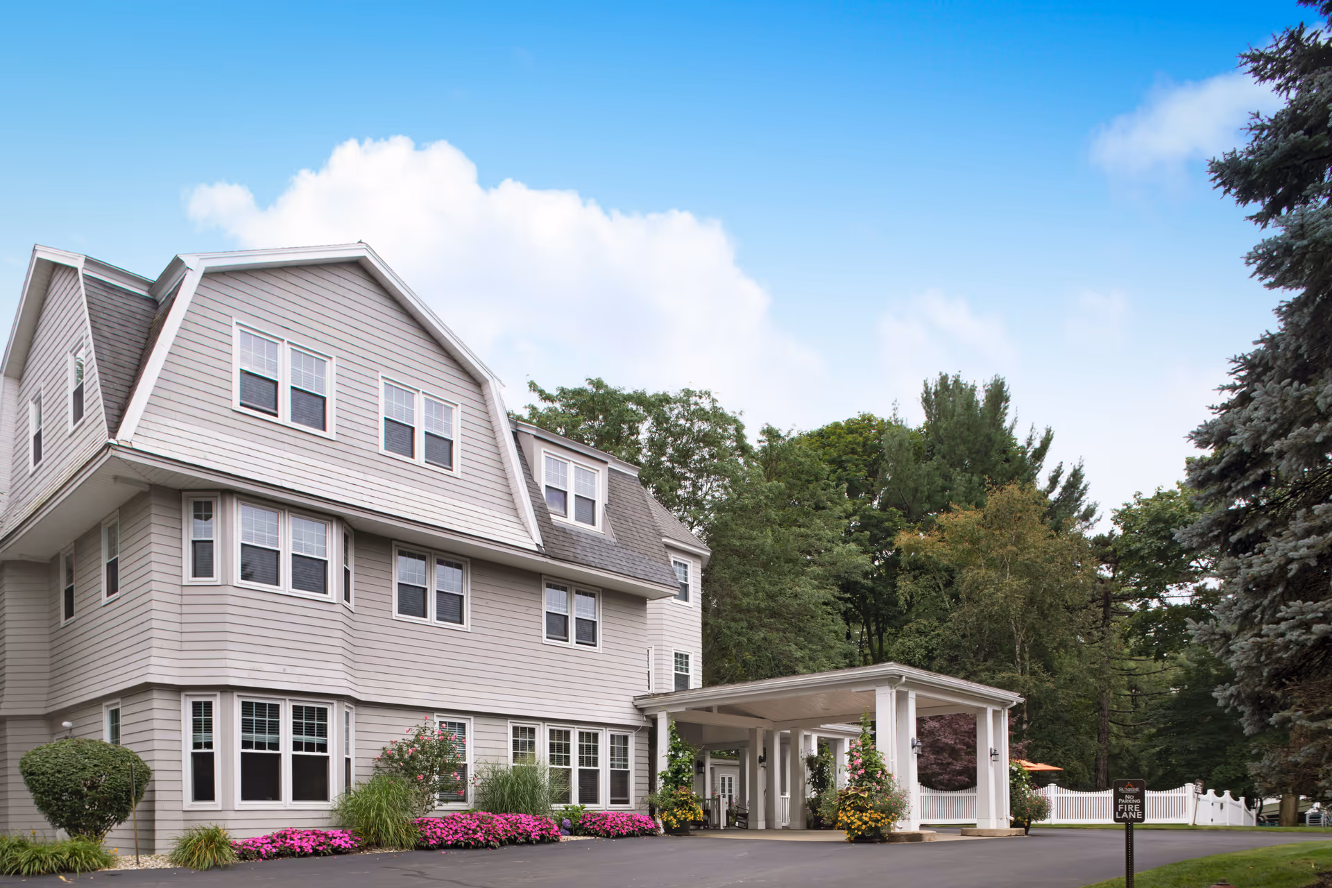 Three-story light gray senior living building with a covered entrance drive, landscaped flower beds, and surrounding trees.