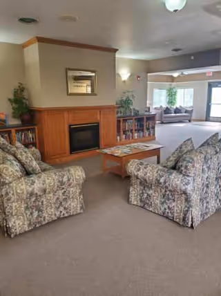 A cozy living room area with two floral-patterned armchairs facing a wooden coffee table and a fireplace with a wooden mantel. Behind the fireplace are bookshelves filled with books and decorative plants. In the background, there is a large window with a sofa and more plants near the entrance door.