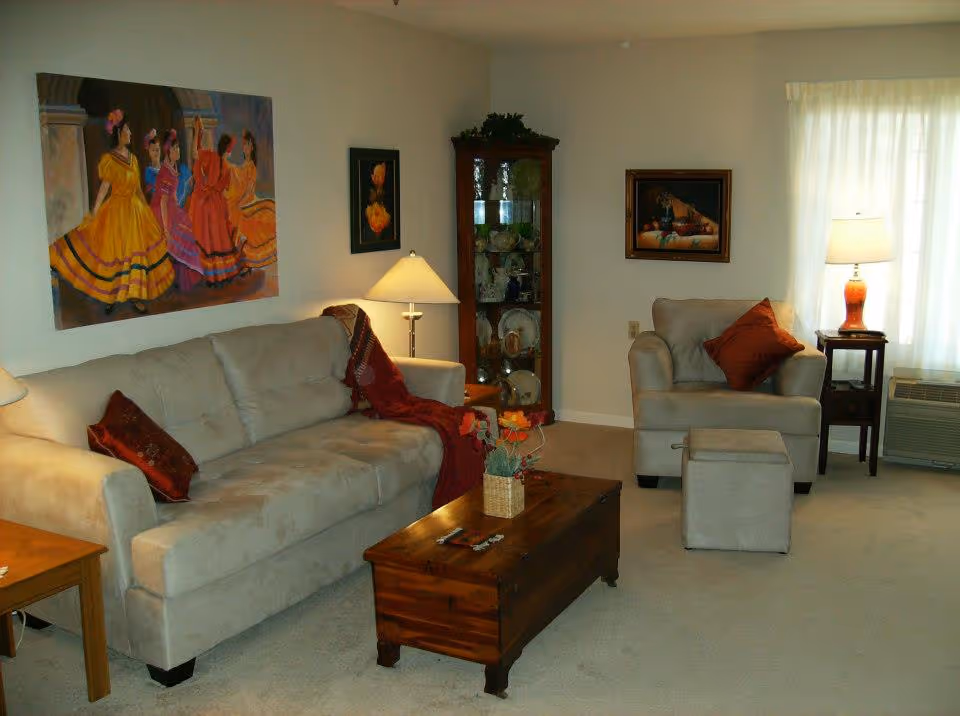 A cozy living room with a beige sofa and matching armchair, both adorned with red accent pillows. A wooden coffee table with a small flower arrangement sits in front of the sofa. The room features a glass display cabinet filled with dishes, two table lamps providing warm lighting, and three paintings on the walls, including one large colorful painting of women in traditional dresses. Light filters in through a window with sheer curtains.
