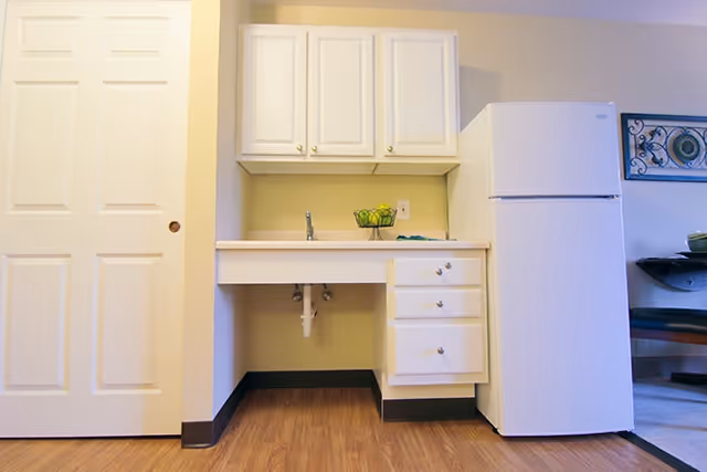 Small kitchenette with white upper and lower cabinets, a sink and drawers, and a white refrigerator next to a sliding door.