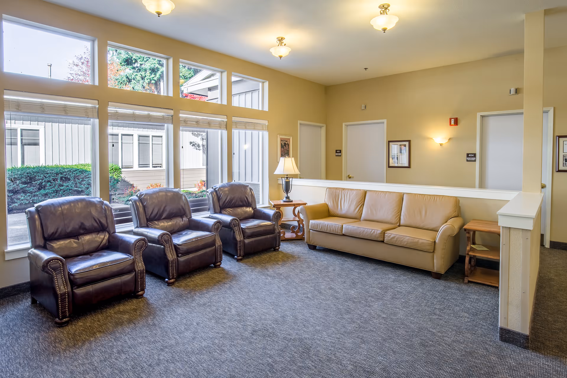 A bright and clean sitting area in a senior living facility with three dark brown leather armchairs and a beige leather sofa arranged around two wooden side tables with a lamp. Large windows let in natural light and show greenery outside. The walls are painted light yellow with framed pictures and two closed white doors labeled 403 and 404.