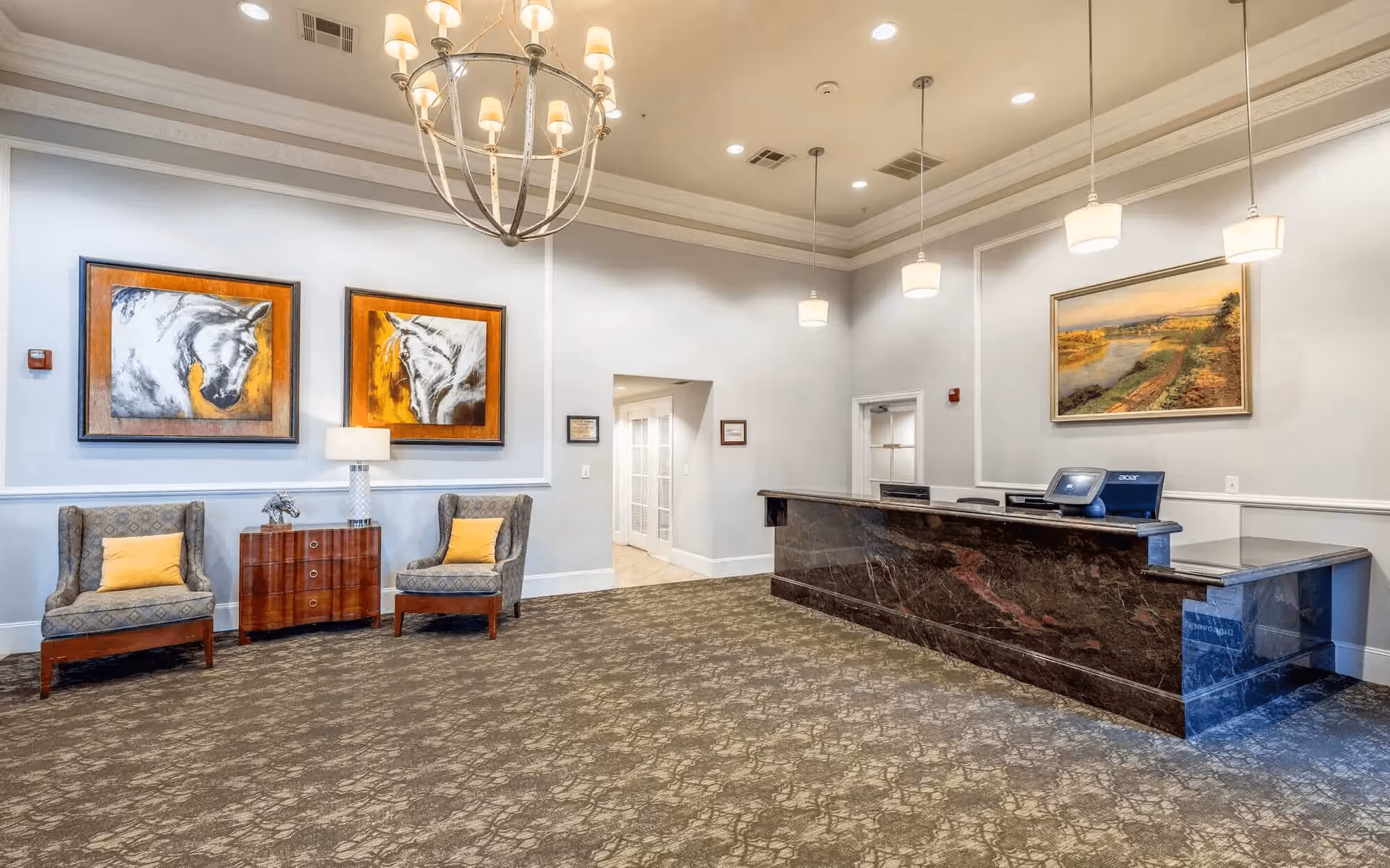 Reception area of Conservatory At Plano featuring a dark marble front desk with computer monitors, two patterned armchairs with yellow cushions, a wooden side table with a lamp and horse sculpture, and framed artwork of horses and a landscape on the walls. The room has a patterned carpet, white walls with molding, and multiple ceiling lights including a chandelier and pendant lights.