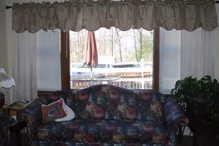 Floral-patterned sofa sits in front of a large window with blinds and a valance, showing a deck and umbrella outside, flanked by side tables and a plant.