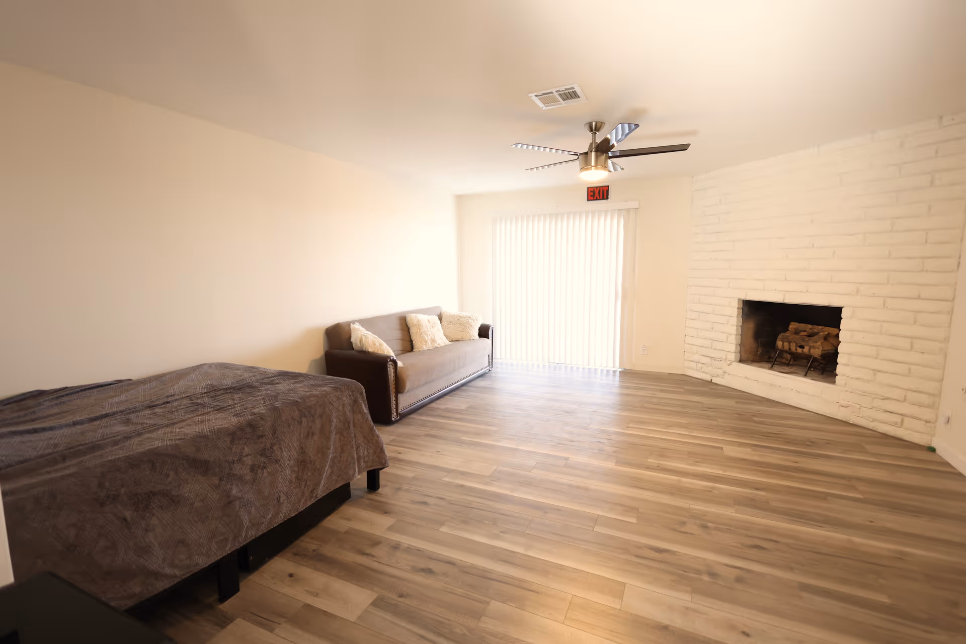 A spacious room with light-colored walls and wood flooring featuring a bed with a dark blanket on the left, a brown sofa with three cushions against the wall, a white brick fireplace on the right, and a ceiling fan with lights. There is a sliding glass door with vertical blinds and an exit sign above it at the far end of the room.