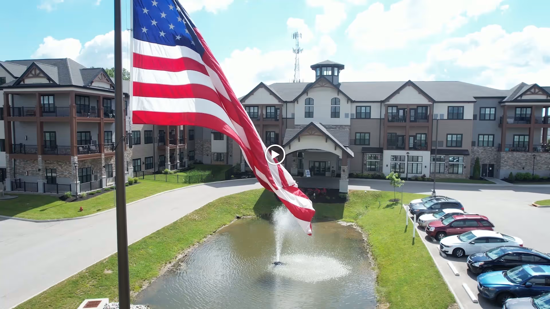 Front exterior of a multi-story senior residence with an American flag flying over a pond fountain and adjacent parking lot.