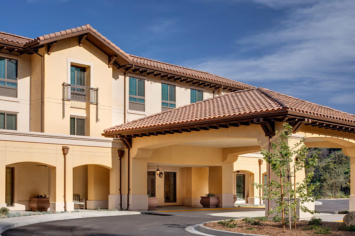 Exterior view of Belmont Village Senior Living Calabasas showing a beige stucco building with a red tile roof, arched entryway, and several windows with green shutters under a clear blue sky.