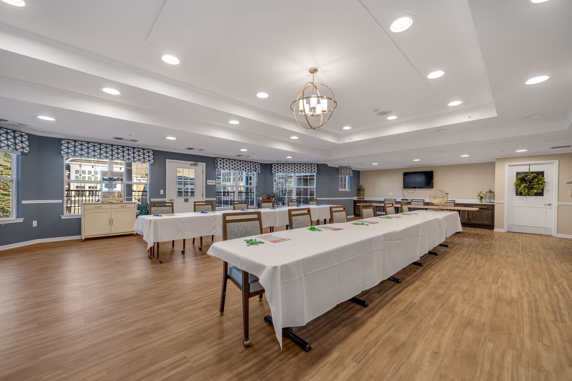 A spacious, well-lit room with long tables covered in white tablecloths arranged in a U-shape. Chairs are placed around the tables, and the room features wood flooring, blue walls with windows covered by patterned valances, and a modern chandelier hanging from the ceiling. A television is mounted on the far wall above a cabinet, and a door with a wreath is visible on the right side.