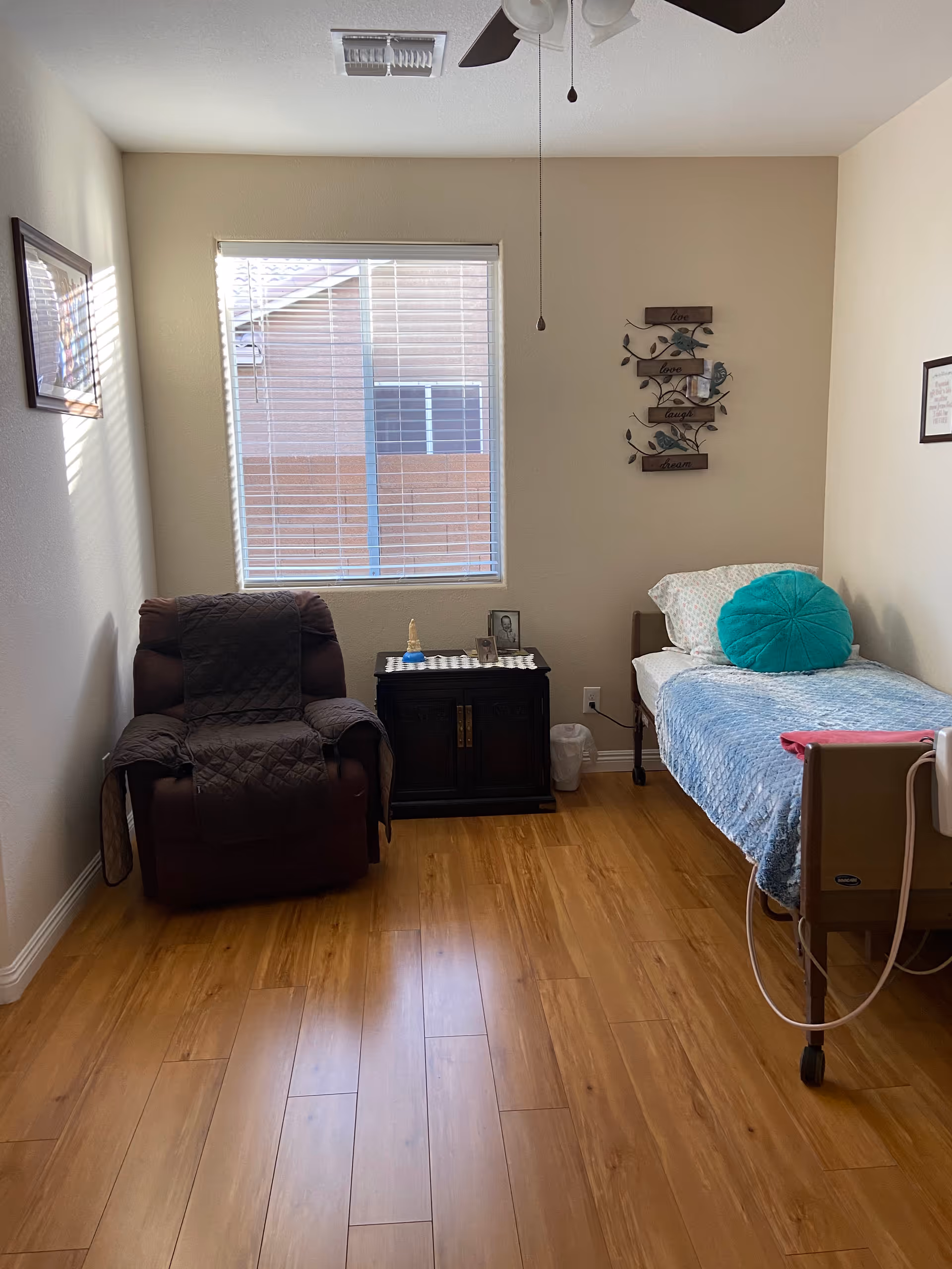 A small, tidy bedroom in an assisted living facility with a single bed on the right side covered with a blue blanket and a round teal pillow. Next to the bed is a small dark wooden cabinet with a few decorative items on top. To the left is a brown recliner chair with a dark quilted cover. A window with blinds is centered on the back wall, allowing natural light to enter the room. The walls are light-colored, and there are framed pictures hanging on both side walls. The floor is wooden, and a ceiling fan with lights is visible above.