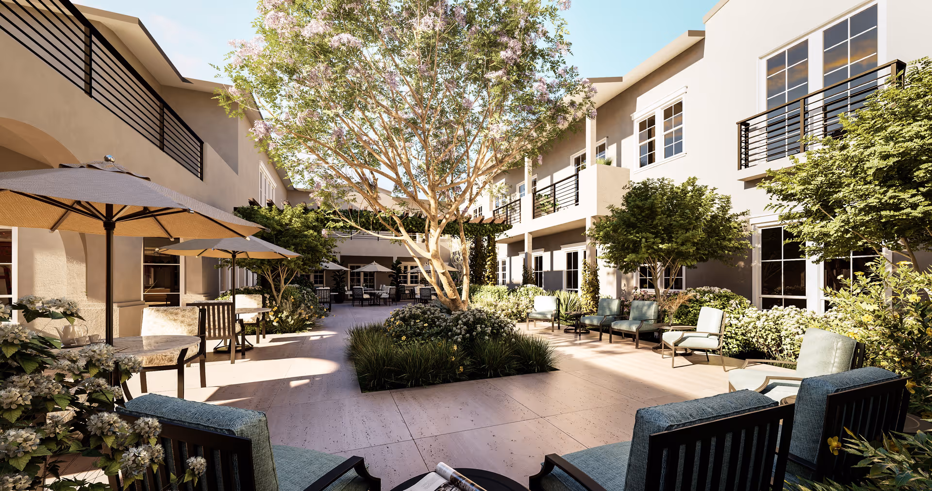 Outdoor courtyard area of a senior living facility with seating arrangements including cushioned chairs and tables with umbrellas. The courtyard is surrounded by a two-story building with balconies and large windows. There are trees, shrubs, and flowering plants providing greenery and shade under a clear blue sky.