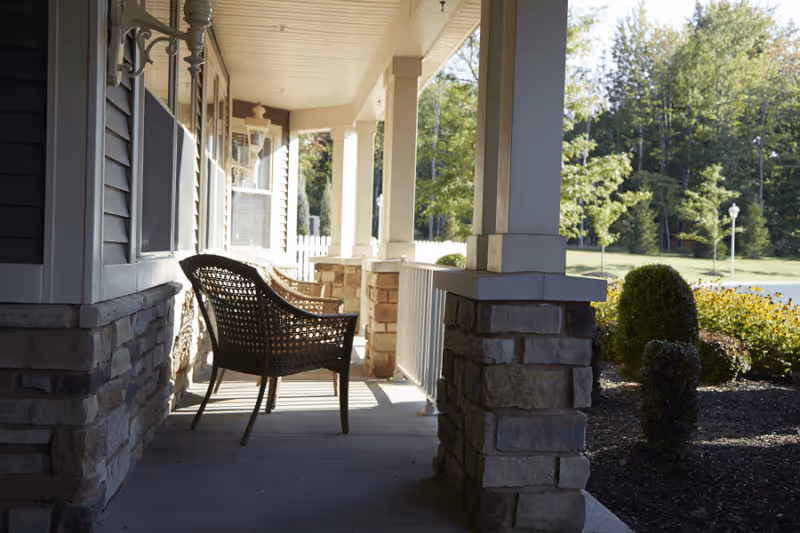Covered front porch with a wicker chair between stone columns overlooking a landscaped lawn and trees.