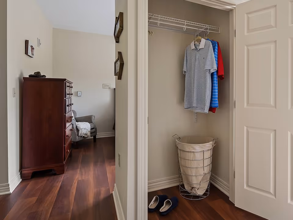 Interior view of a room featuring an open closet with three shirts hanging on a wire shelf and a laundry basket below. On the floor near the closet are a pair of slippers. To the left, a wooden chest of drawers and a cushioned chair with a blanket draped over it are visible. The room has wooden flooring and light-colored walls.