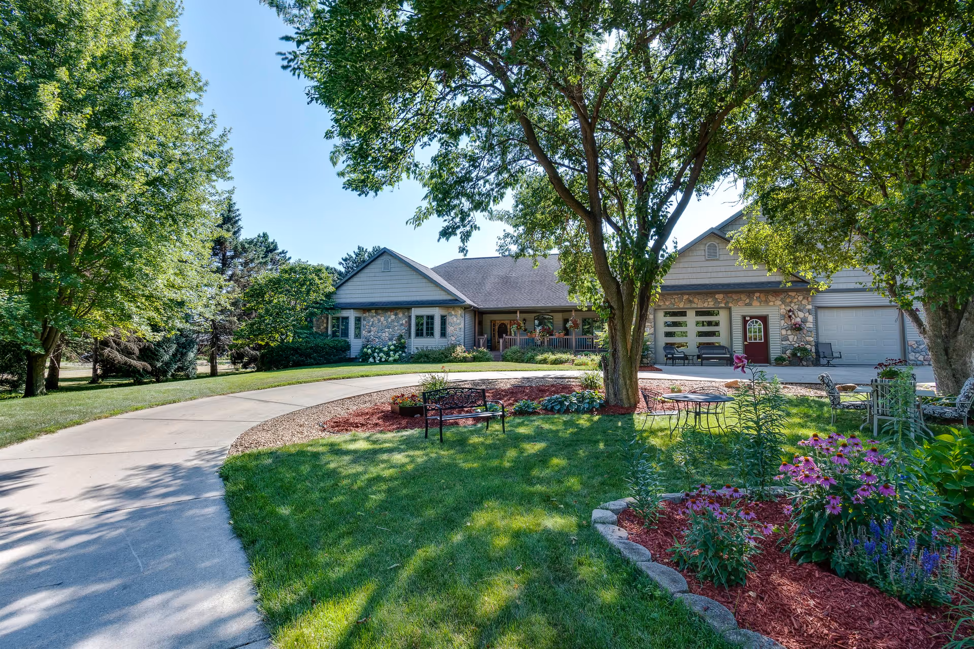 A single-story senior living facility building with a stone and siding exterior, surrounded by lush green trees and a well-maintained garden with colorful flowers. There is a curved concrete driveway leading to the entrance, outdoor seating areas with benches and chairs, and a bright blue sky overhead.