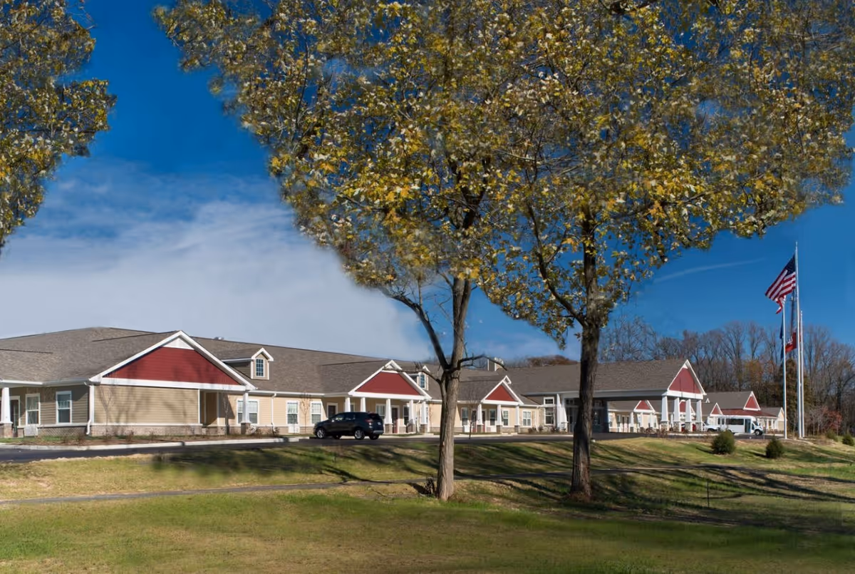 Exterior view of a single-story senior living facility building with beige and red siding, multiple windows, and a covered entrance. There are two trees in the foreground, a grassy lawn, and three flagpoles with flags flying on the right side of the image under a clear blue sky.