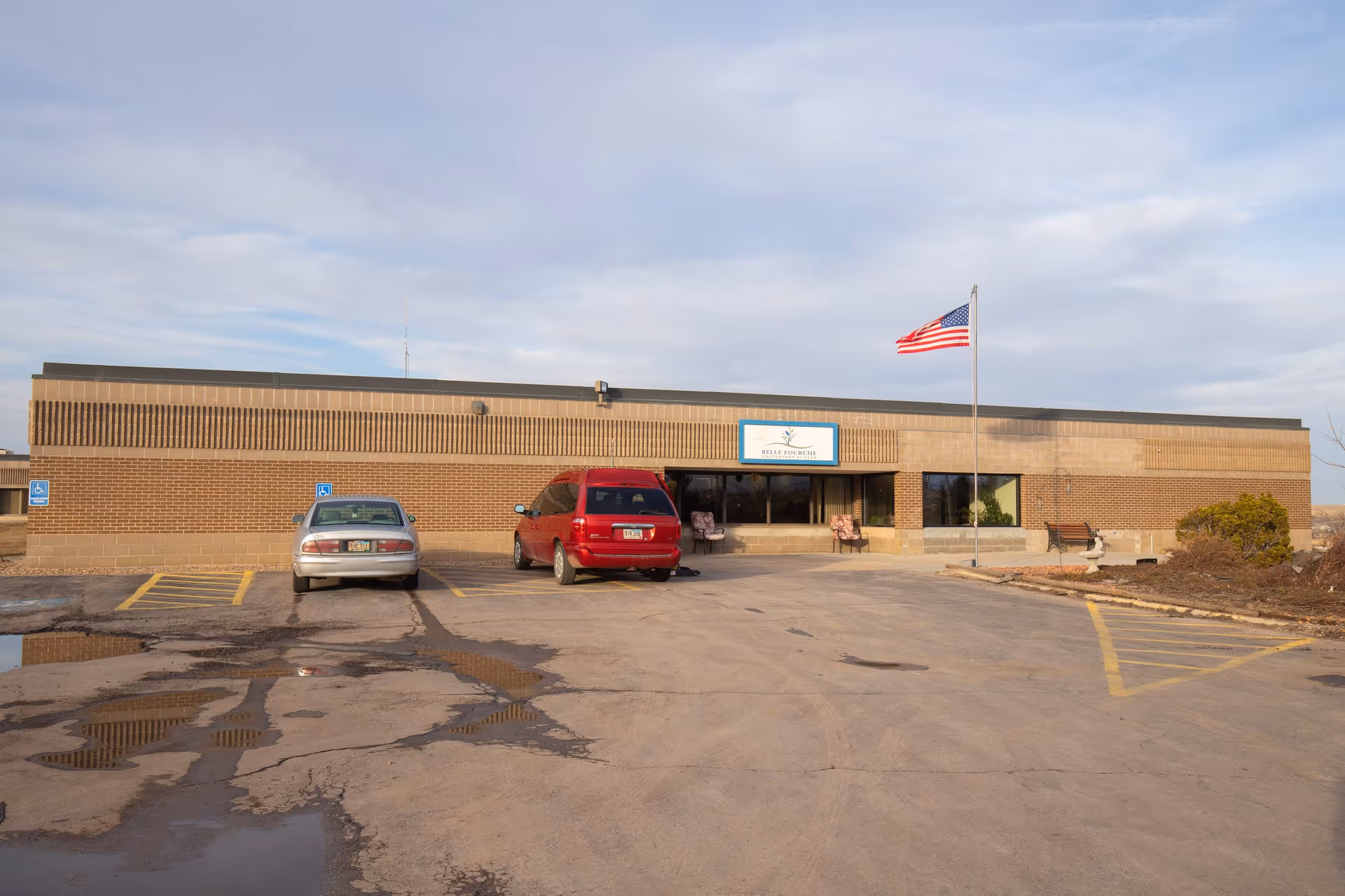 Exterior view of a single-story brick building with a flat roof, two parked cars in front, an American flag on a flagpole, and a sign above the entrance reading 'Rolling Hills Healthcare'. The parking lot has some puddles and handicap parking spaces.