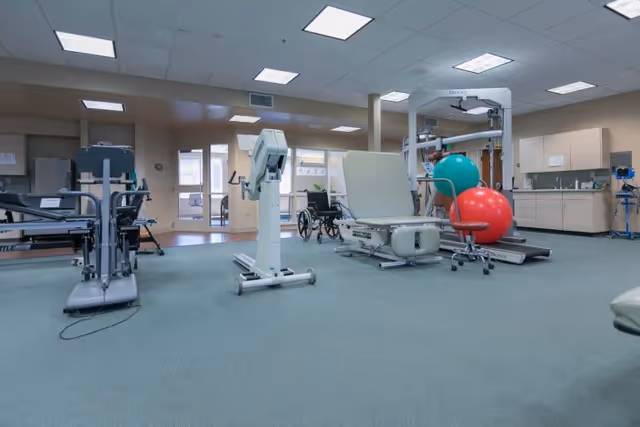 Therapy gym with exercise machines, a treatment table, large red and green therapy balls, and a wheelchair.
