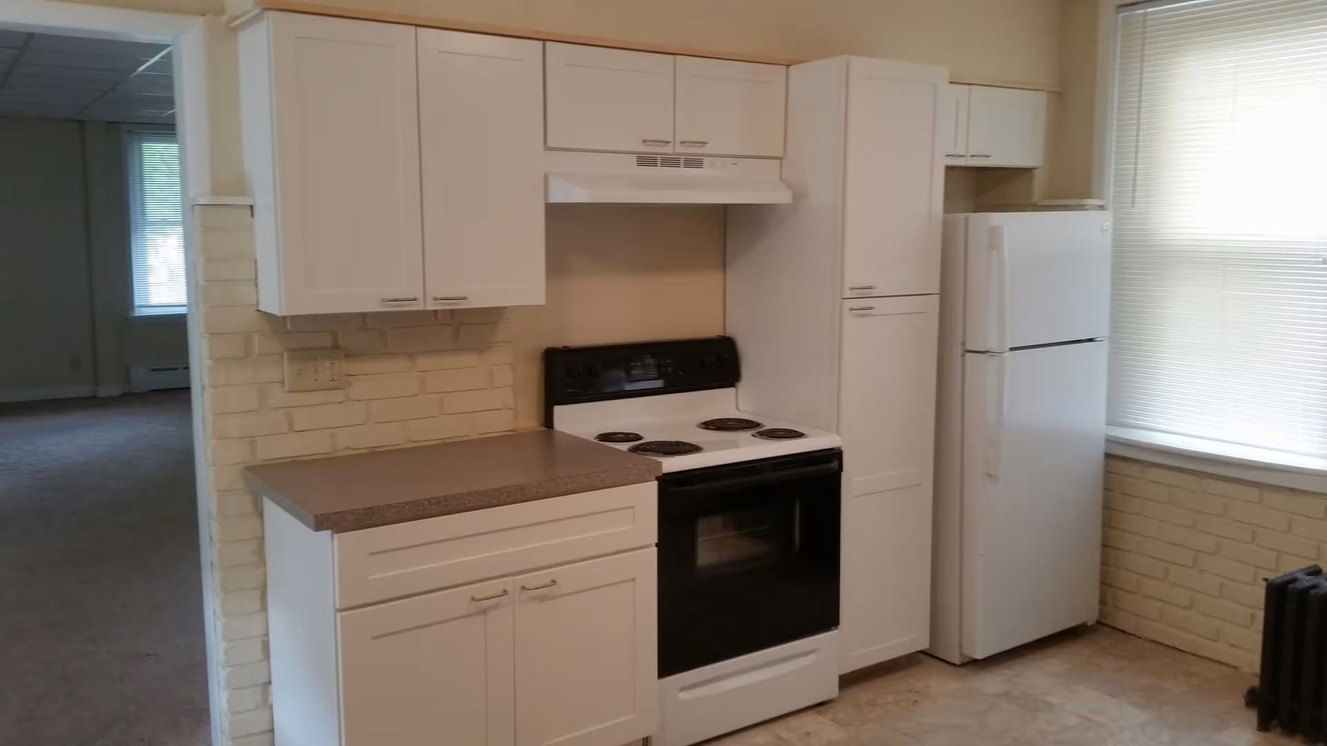 A kitchen area with white cabinets, a white electric stove with four burners, a white refrigerator, and a countertop. The walls have a light brick pattern, and there is a window with blinds letting in natural light.